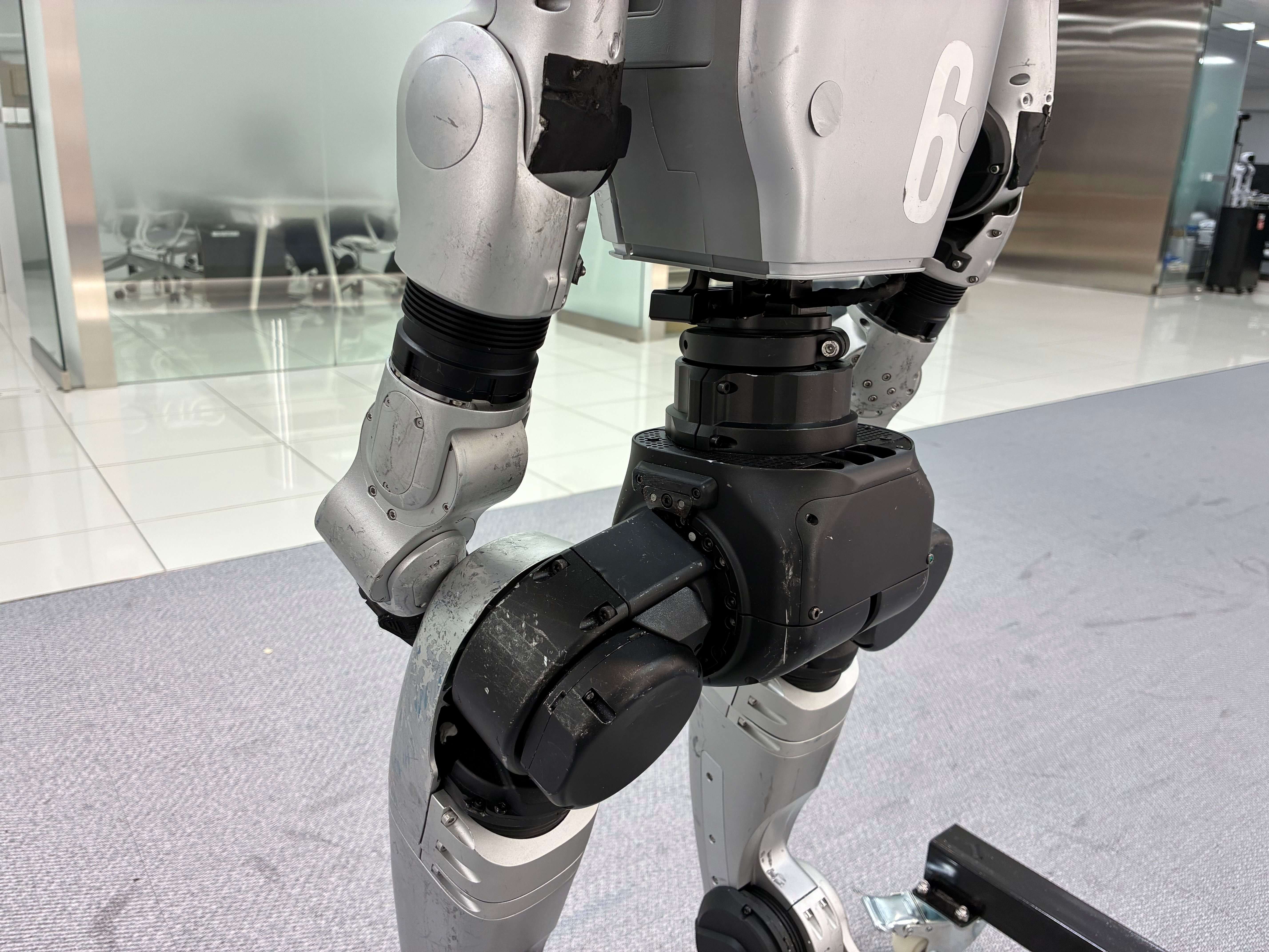 ヒューマノイド（人型ロボット）の腰部から脚部にかけてを間近で捉えたクローズアップで、白い数字「6」が記された擦れの目立つシルバー外装、蛇腹状ブラックカバーで覆われた関節部、ボルトや通気スリットが並ぶ黒い腰部ユニット、体側に沿って下ろされた腕部などの駆動機構が細かく写り、背景にはガラス張りの会議室とタイル床のオフィスがぼかしで広がっている画像。