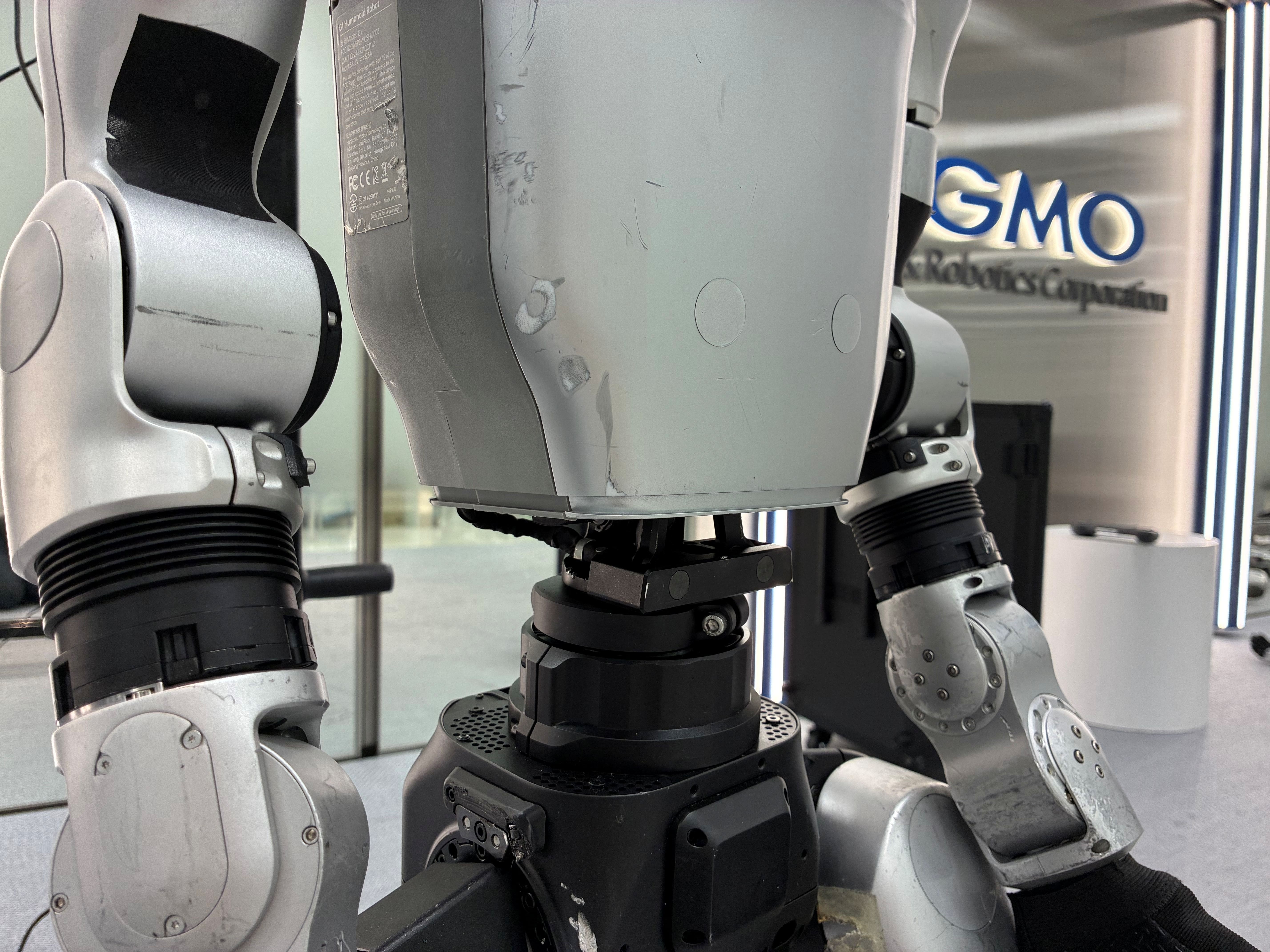 Unitree製ヒューマノイド（人型ロボット）の胴体下部から脚部にかけてを間近で捉えたクローズアップで、使用による擦れや傷が残るシルバーの外装パネル、蛇腹状のブラックカバーに覆われた股関節、ボルトが並ぶ黒い腰部ユニットや通気孔、側面の認証マークラベルなどメカ構造が細かく写り、背景には「GMO AI&Robotics Corporation」のロゴが入ったオフィスの壁面がぼんやりと見える画像。