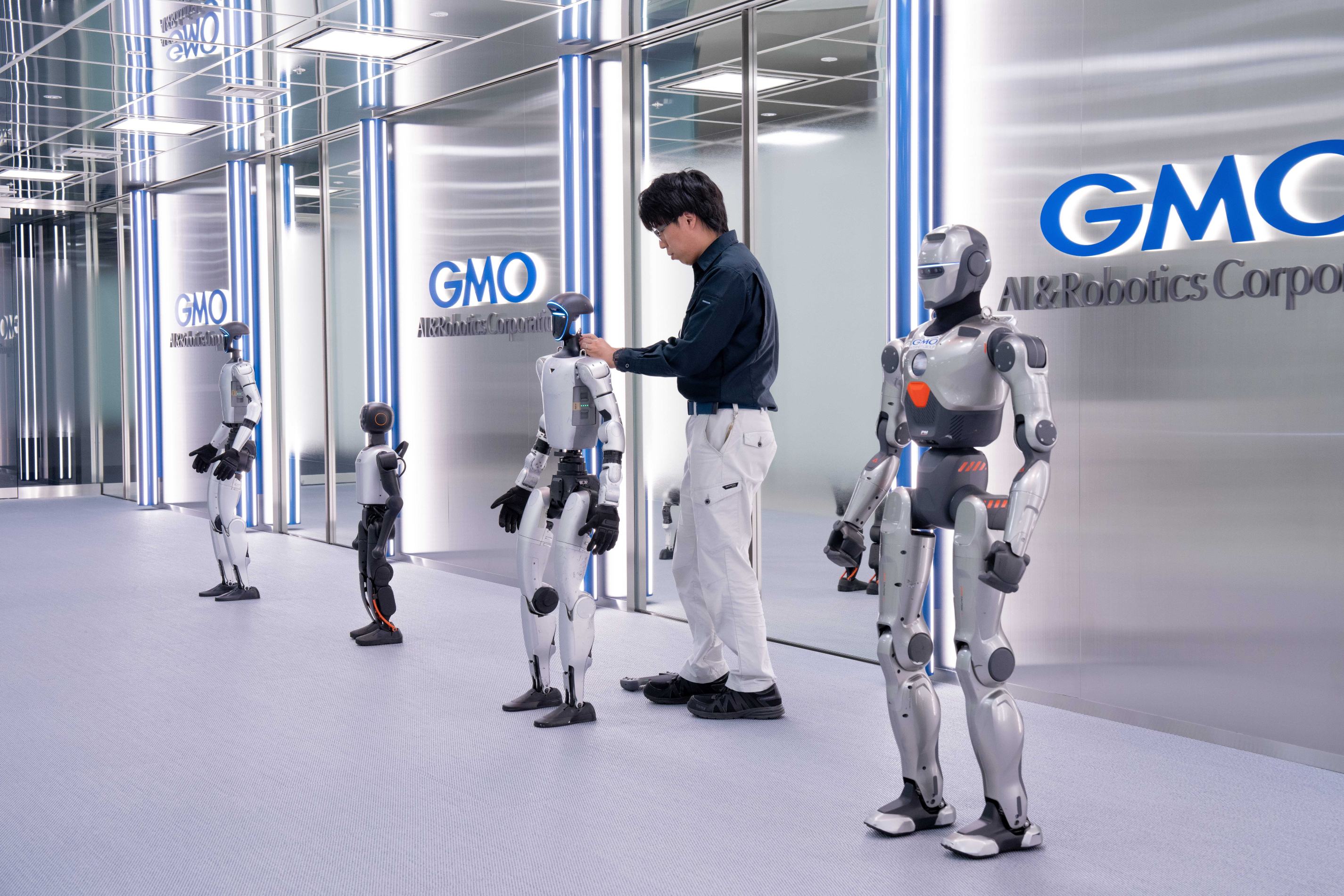 GMO AI＆ロボティクス商事のオフィスと思われるメタリックな内装の空間で、壁面にブルーの「GMO AI&Robotics Corporation」ロゴが並ぶ通路に4体のヒューマノイド（人型ロボット）が立ち並び、中央では眼鏡をかけた黒シャツ・白作業ズボン姿の男性スタッフが1体の頭部付近に手を添えて調整・点検を行っており、右手前にはオレンジのアクセントが入ったシルバーの大型機体が正面を向いて直立している画像。